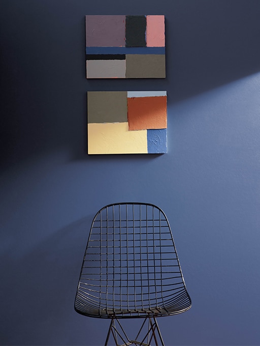 A deep blue wall featuring art that represents the eight colours of the Benjamin Moore Colour of the Year colour palette and a black metal basket chair