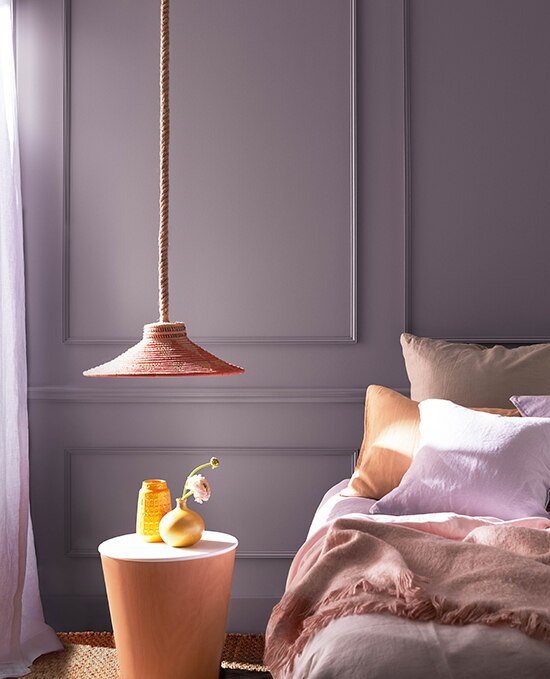 A panelled bedroom painted in Hazy Lilac 2116-40. The styling features neutral texiles, a pendant lamp and a bedside table decorated with golden vases