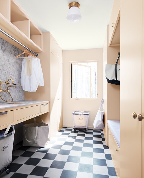 A laundry room showcasing cabinets painted in Pristine OC-75.  Styling features a checkerboard tile, gold taps, marble work-surface and fabric laundry baskets