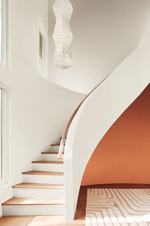 A curved staircase painted in White Dove OC-17 featuring a bold Topaz 070 accent wall beneath it. Styling includes a neutral area rug and a rice paper chandelier