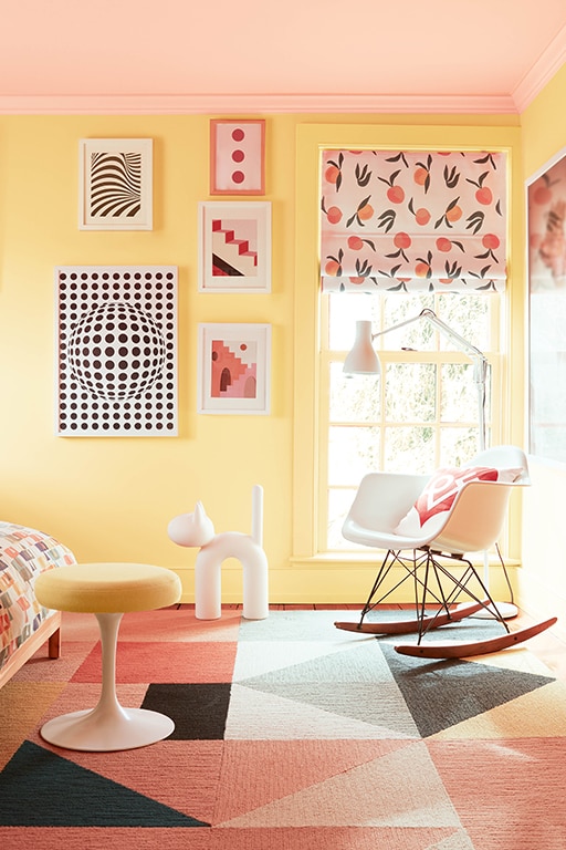 A playful bedroom with a ceiling pained in Teacup Rose 2170-50 and walls painted in Honeybee CSP-950. Styling includes a rocking chair, pattered blinds and bold art