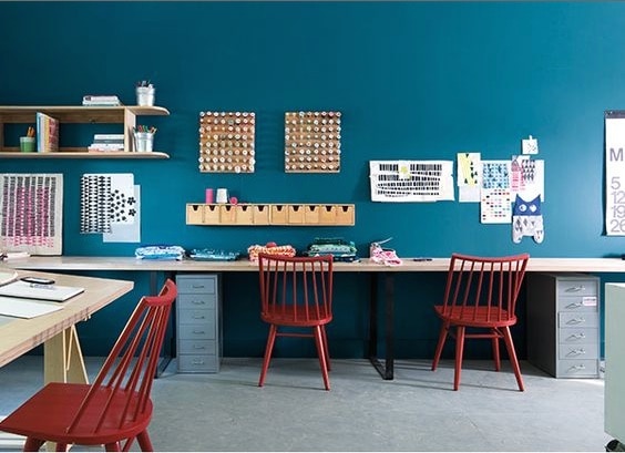 A communal workspace with walls painted in the vibrant Avalon Teal CSP-645. Three rich red chairs are placed along a long wooden work surface, which is decorated with office and craft materials